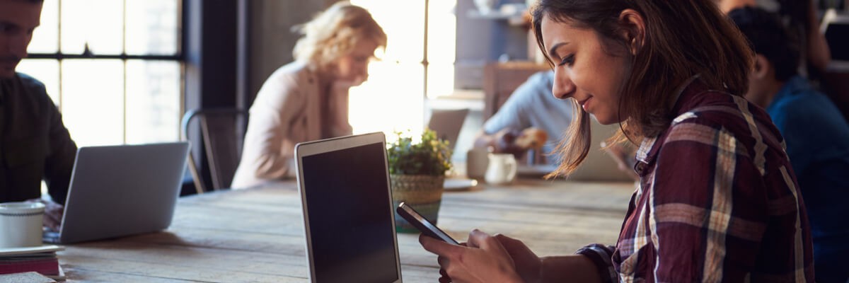 What Small Businesses Need to Know about BYOD Security A girl sits at a cafe and looks at her phone while her laptop is also open on the table.